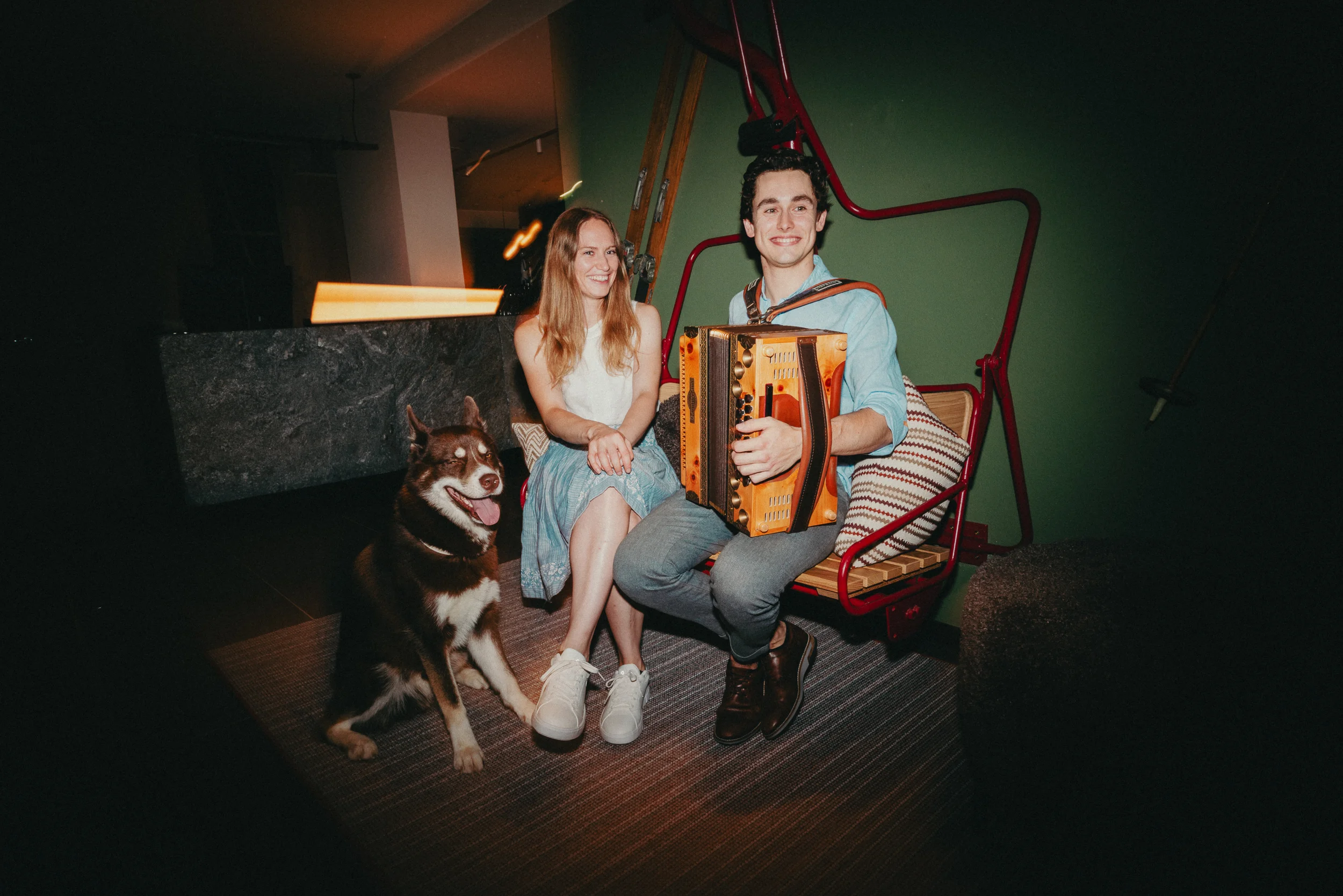 Die Gastgeber Daniela und Florian sitzen auf einem Sofa zusammen und Florian spielt Zieharmonika